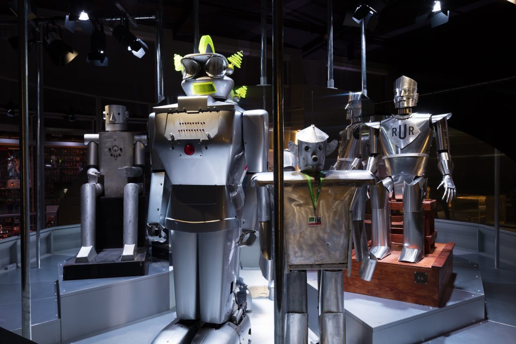 A number of historic-looking silver robots with boxy features sit and stand in an exhibition. 