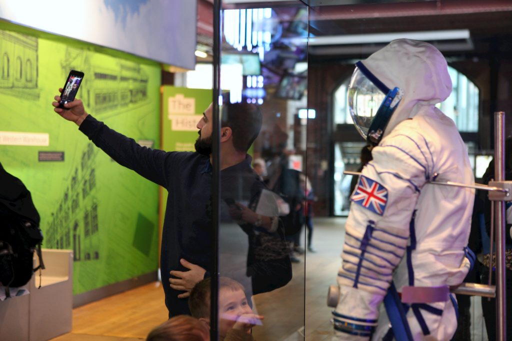 A man takes a selfie in front of Tim Peake's spacesuit, a white astronaut suit with a Union Jack badge on the sleeve and a clear-fronted helmet