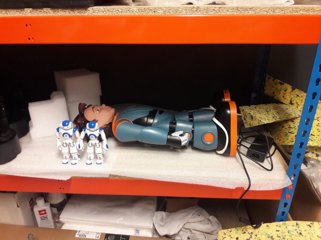 A humanoid robot resembling a small boy lies on a trolley alongside two smaller blue and white robots