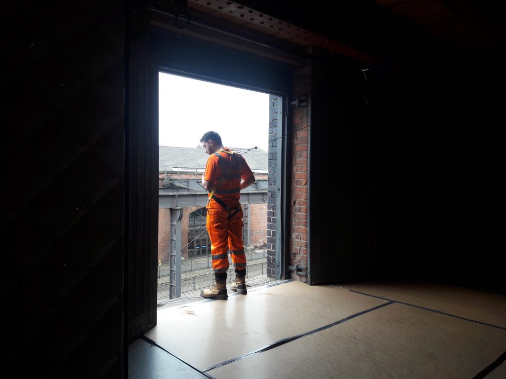 A worker in an orange uniform and harness watches the deinstall process from the opening in the gallery where objects are removed