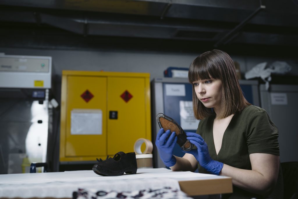 A woman with brown hear wearing a green dress and blue gloves sits in a museum room inspecting a small pair of leather clogs
