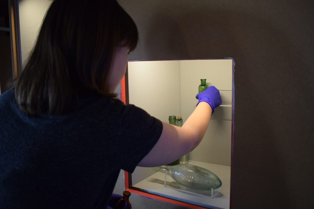 A woman wearing blue gloves places a green glass bottle into a display case during the textiles gallery refresh