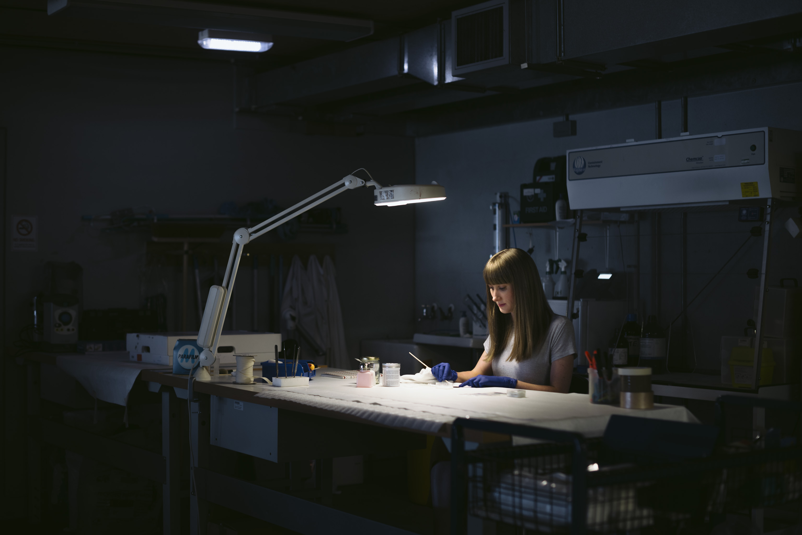 A woman wearing blue gloves sits at a desk in a basement conservation studio. She is concentrating on cleaning a small object in front of her. Her face is dramatically lit from above by a desk lamp. 