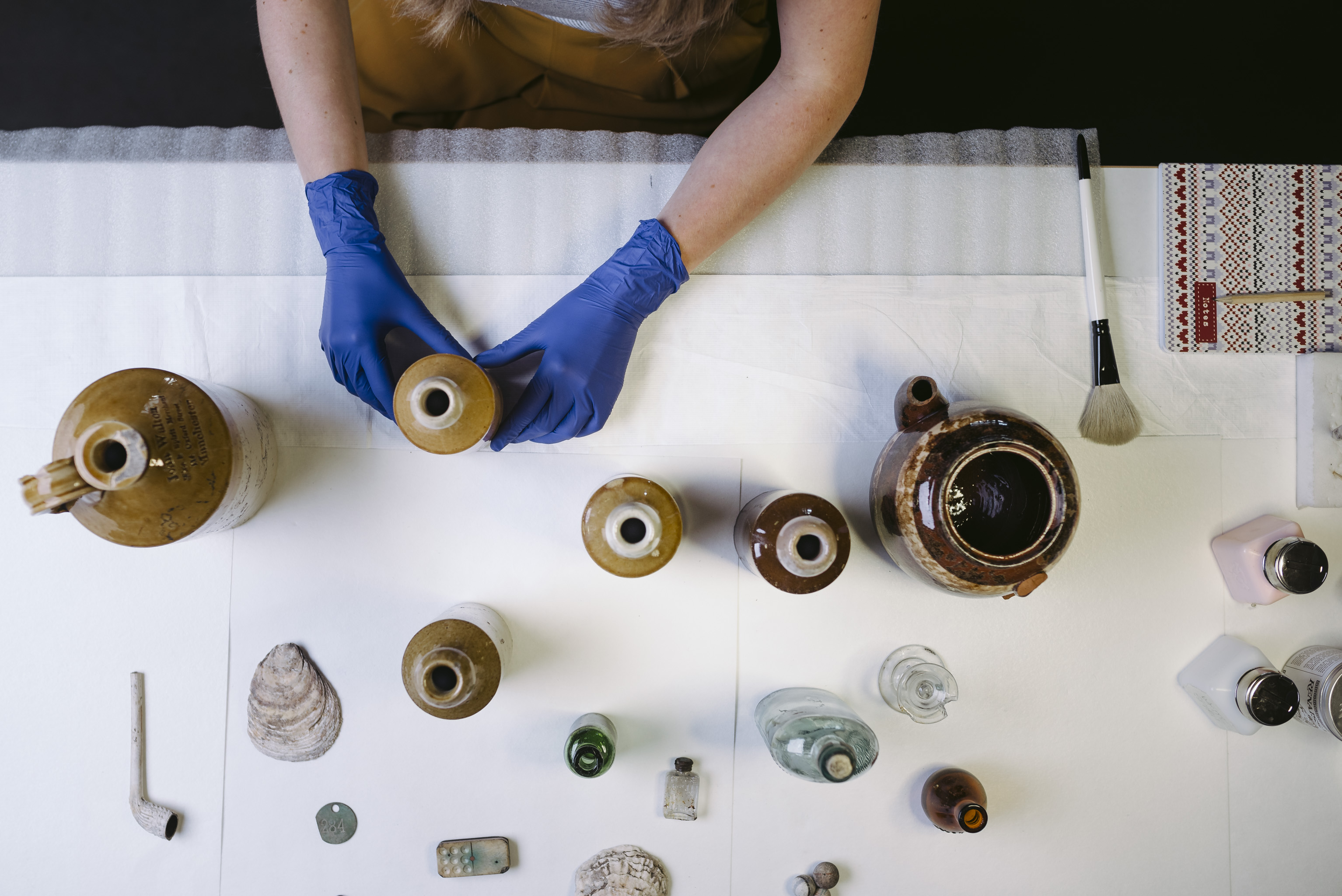 A woman wearing blue gloves handles a selection of pots, bottles and shells