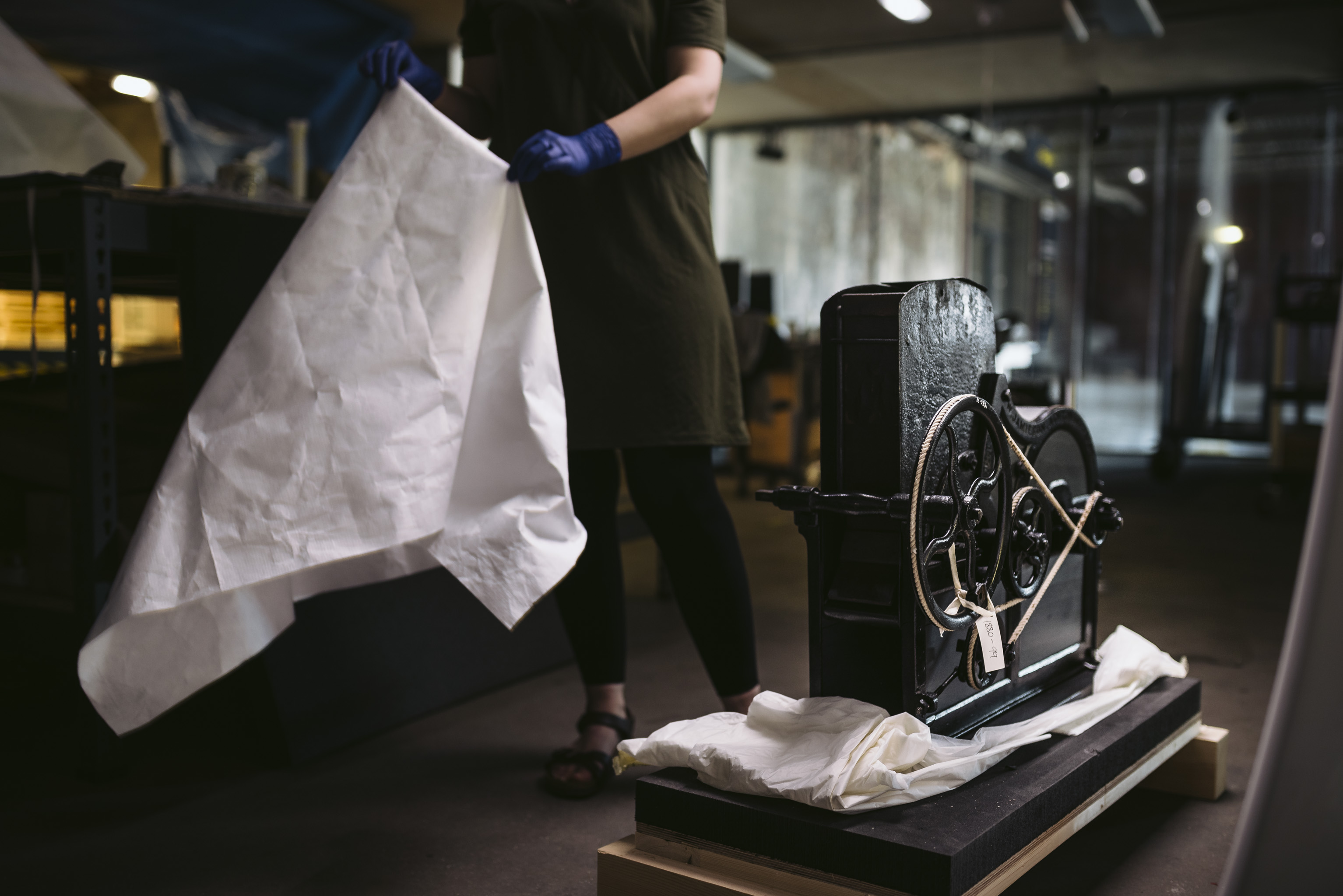 A woman pulls a white covering sheet of a piece of black machinery with a wheel and ropes on one side. 