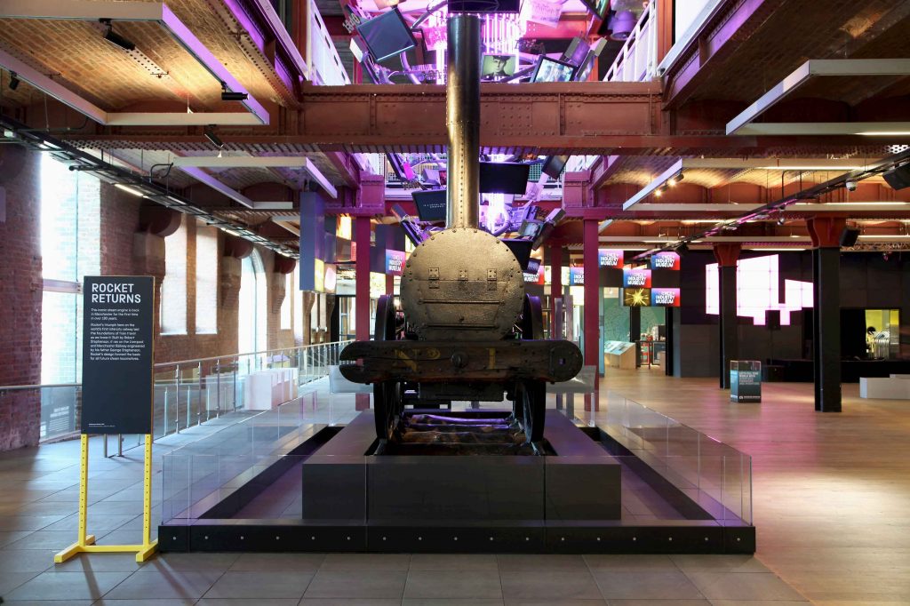 An old fashioned locomotive with a circular "face" and tall chimney, on display in the Revolution Manchester gallery at the Science and Industry Museum in Manchester