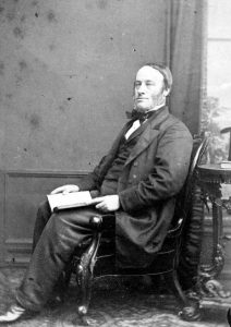 A black and white photograph of a James Joule, a middle aged man with sideburns wearing a formal suit and holding a book on his lap. 