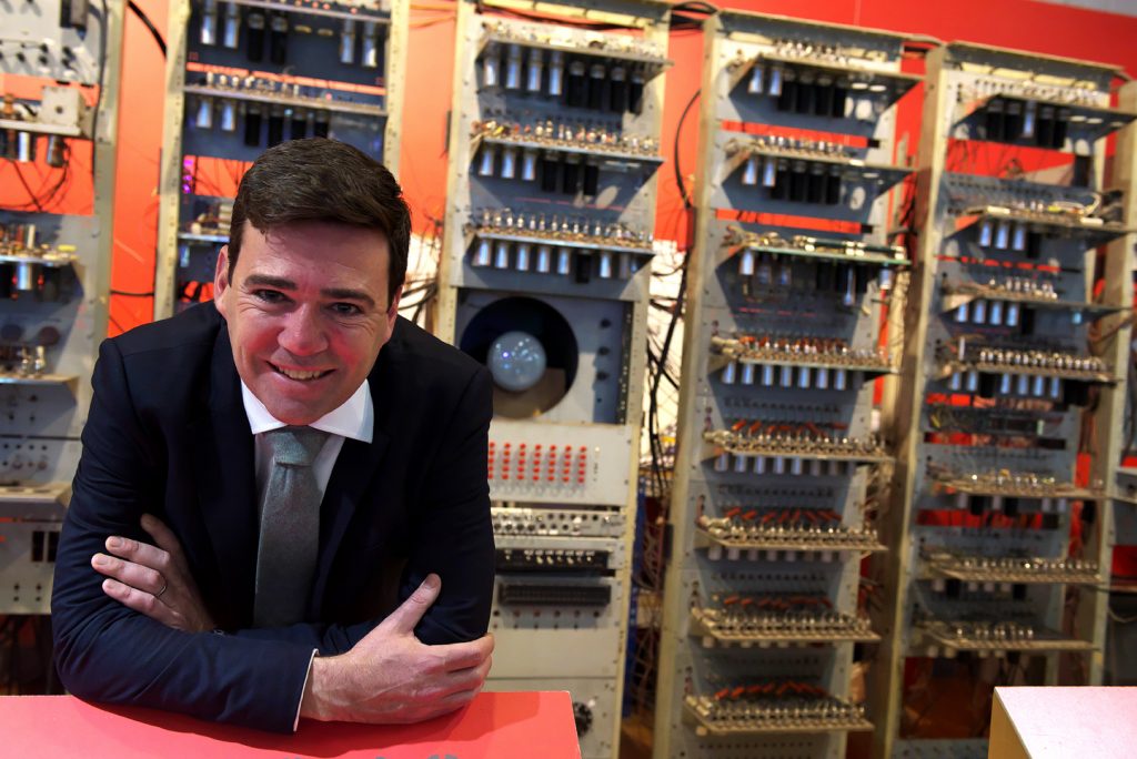 Mayor of Manchester Andy Burnham - a man with dark hair and a navy suit - poses in front of the Small Scale Experimental Machine, a collection of valves and wires that forms the world's first stored-programme computer. 