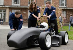 Women in engineering profile subject Pippi Carter-Hornsby with the Formula Student racing team. Three students in blue uniforms, including Pippi (centre) push a black racing car driven by another student wearing a white crash helmet. 