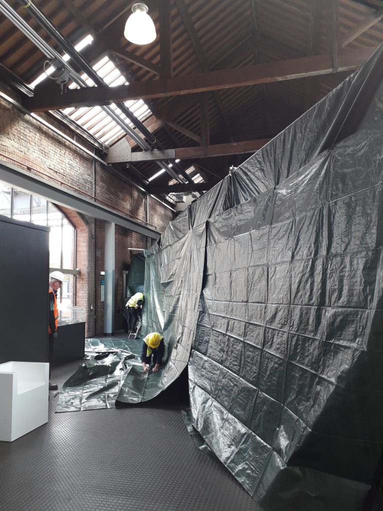 Three people in hard hats and hi-vis vests work to cover a huge machine in green tarpaulin at the Science and Industry Museum's Power Hall