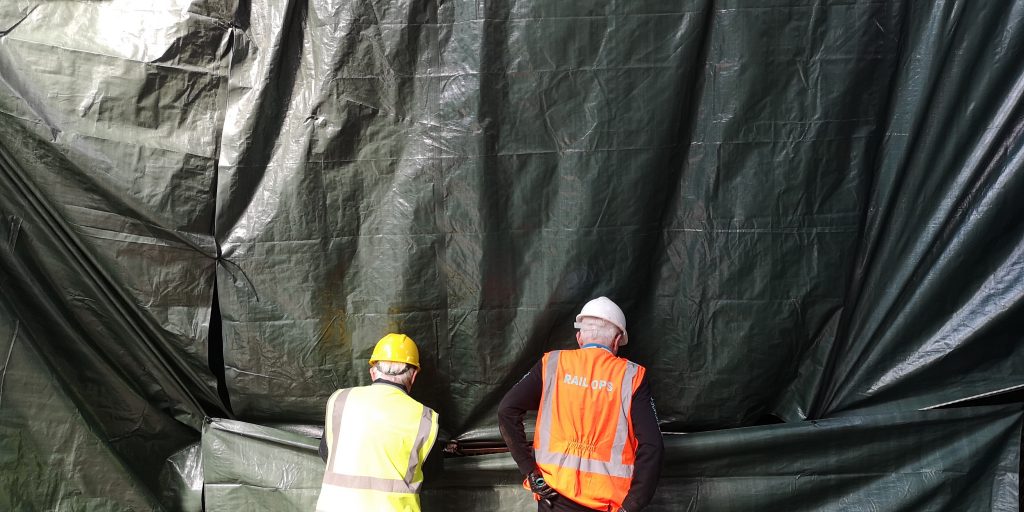 Two men wearing hard hats and hi-vis vests stand in front of a huge sheet of tarpaulin at the Science and Industry Museum's Power Hall