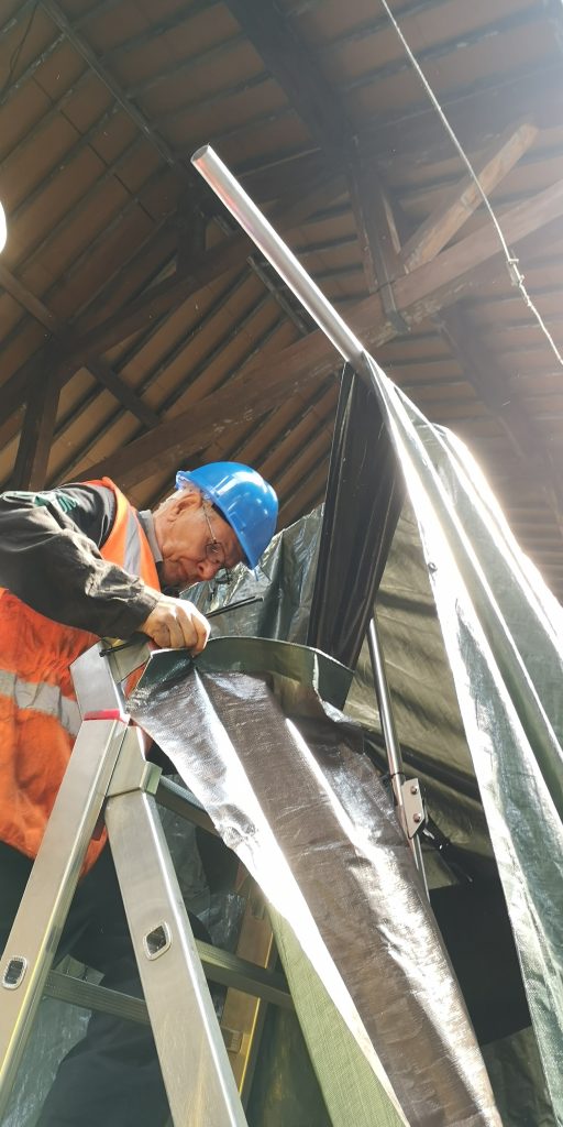 A man wearing a blue hard hat and yellow hi-vis vest stands at the top of a ladder fixing two tarpaulins together in the Science and Industry Museum's Power Hall