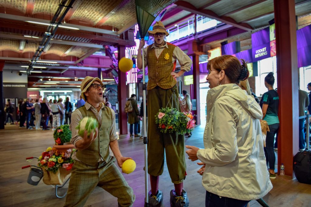 Two men dressed in old-fashioned gardening gear, one is juggling melons, the other is on stilts with a very large rake. Part of the summer of fun at the Science and Industry Museum