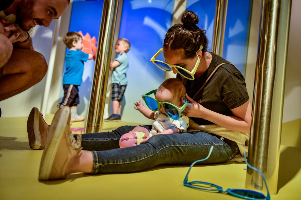 A woman and baby sit on the indoor beach in the Sun exhibition at the Science and Industry Museum. Both are wearing giant sunglasses. 