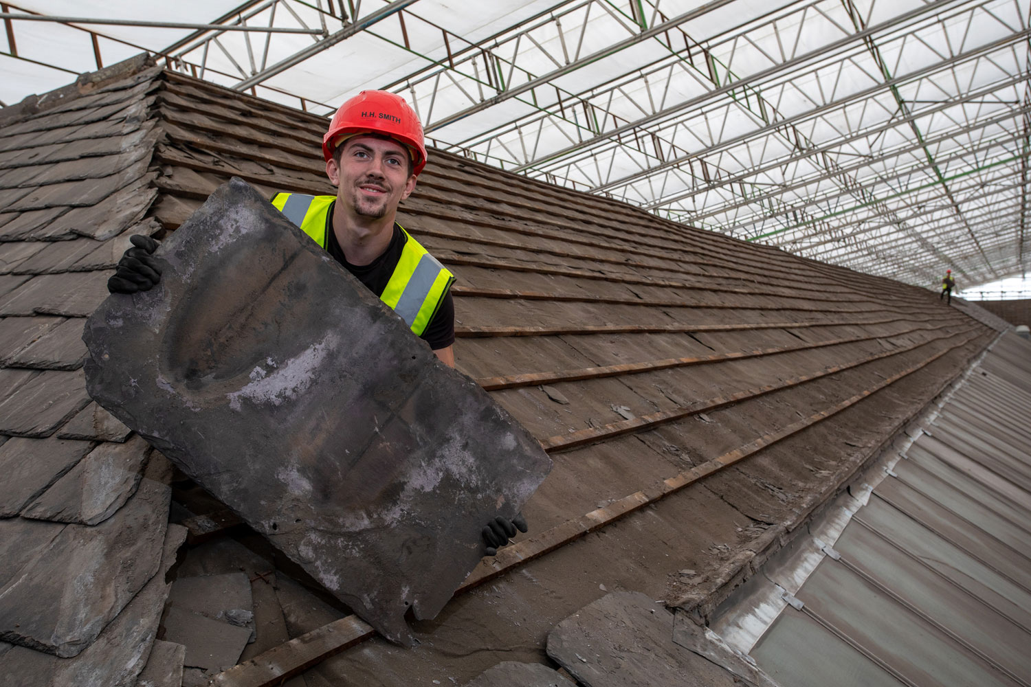 A man on a roof wearing a high-vis jacket and hard hat, holding a large slate roof tile