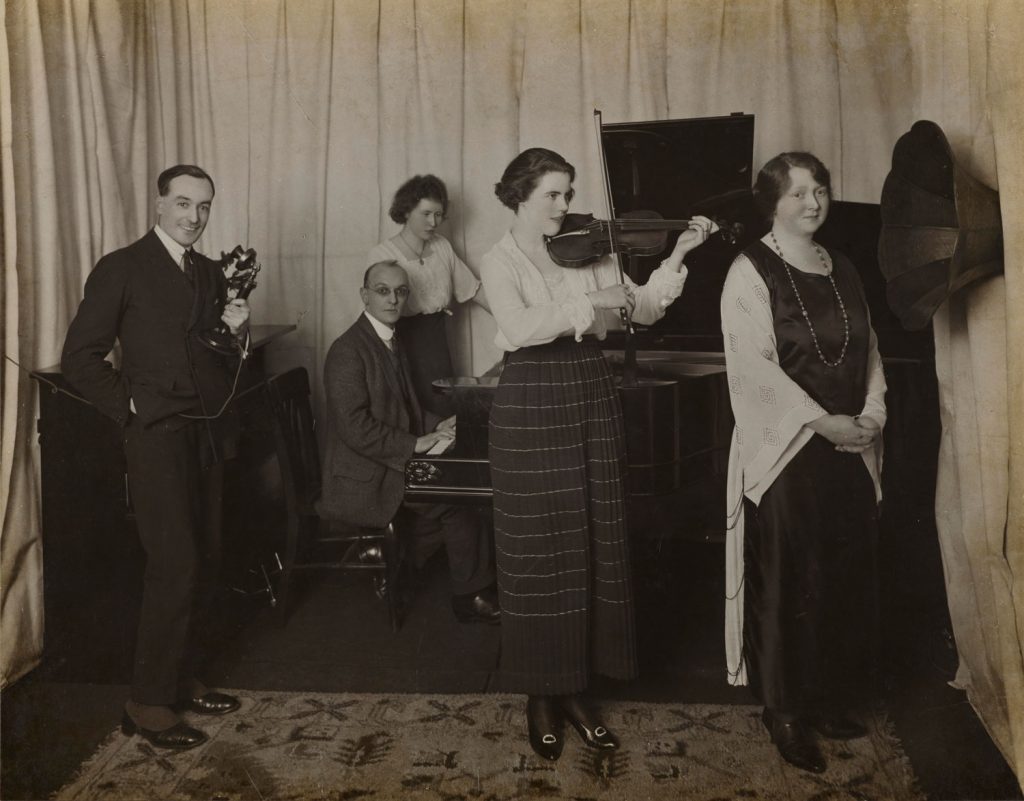 Sepia photo of two men and three women singing and playing musical instruments