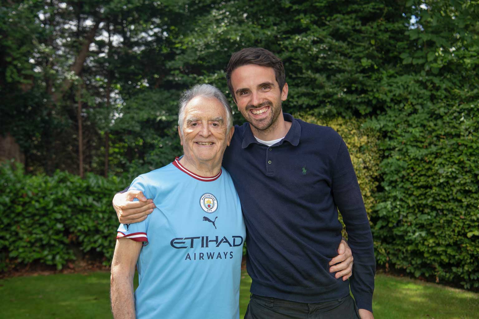 An older man wearing a Manchester City shirt posing for a photo with his adult grandson