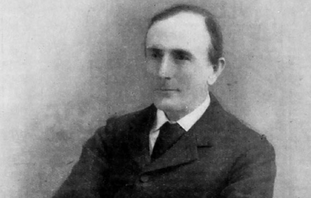 A black and white photo of a man with dark receding hair, wearing a suit and tie 