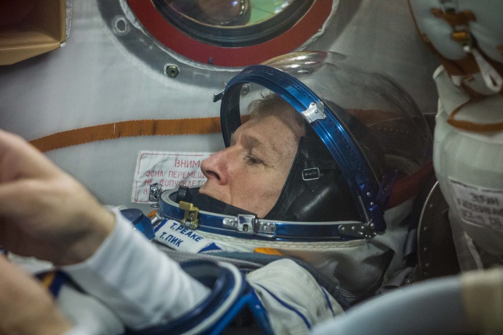 Astronaut Tim Peake checks that his spacesuit fits ahead of his journey to the International Space Station. He lies on his back with the helmet visor open while technicians test the fit. 