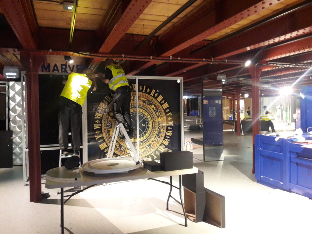 Two workers in hi-vis jackets stand on ladders to move a large and obviously old clock face