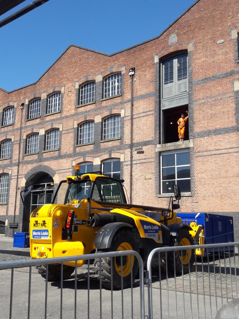 A worker wearing a harness stands in a window high off the ground, watching a large yellow telehandler machine, similar to a tractor, in action