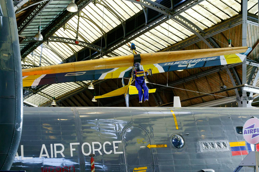 A hang glider with a mannequin steering it, hanging from a steel and glass roof 