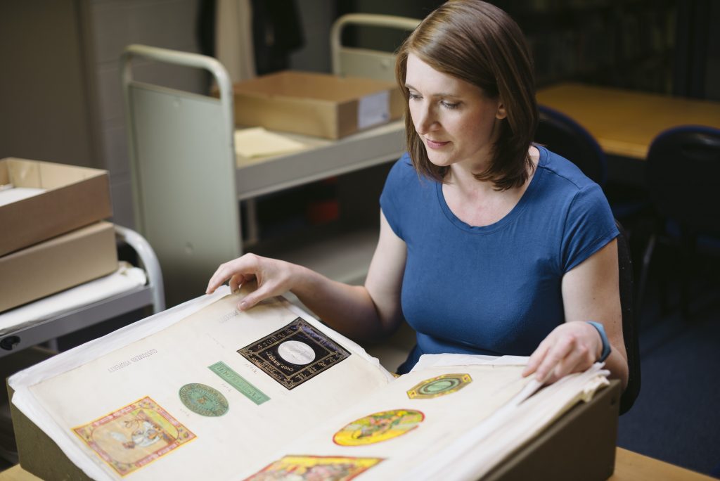 A woman with brown hair and a blue top leafs through a large album containing brightly coloured shipper's tickets