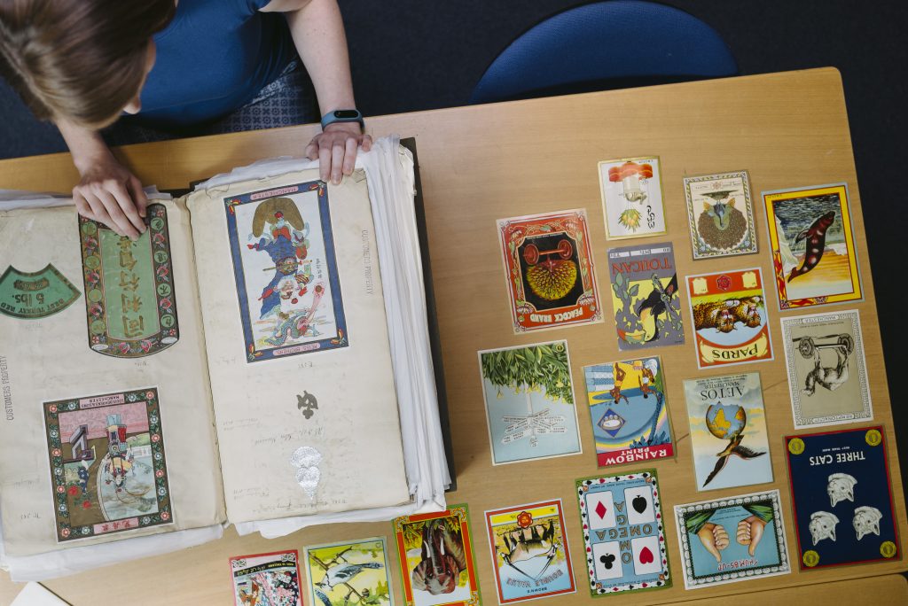 A woman wearing a blue top leafs through a very large album containing colourful shipper's tickets. A selection of tickets are displayed on the table next to her. 