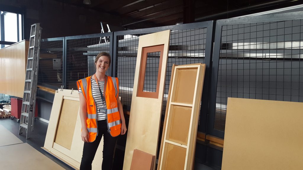 A woman in an orange hi-vis vest stands next to some wooden doors and shelves