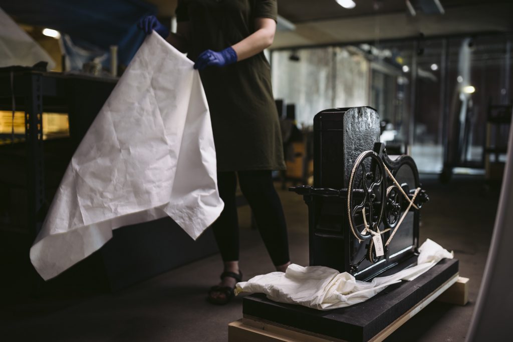 A woman pulls a white covering sheet of a piece of black machinery with a wheel and ropes on one side. 