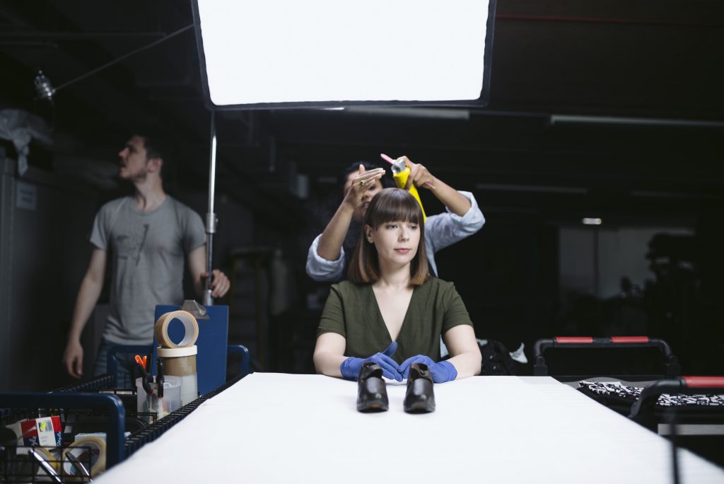 A woman sprays setting spray onto another woman's hair, while in the background a photographer adjusts some lights. 