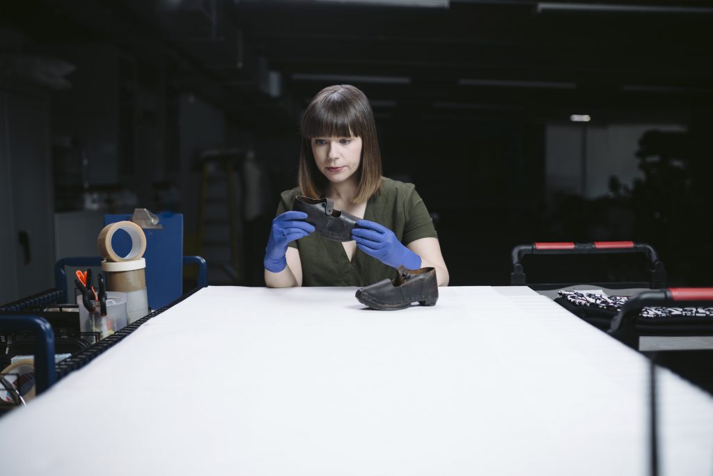 A female curator looks intently at a small pair of leather clogs. She is seated at a large desk surrounded by shelves and trolleys. 