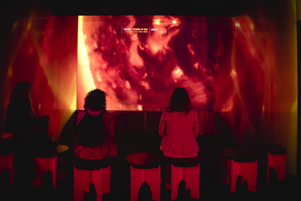 Two women watch a screen on which there is a close up of the Sun's surface, a dramatically firey orange and black swirl