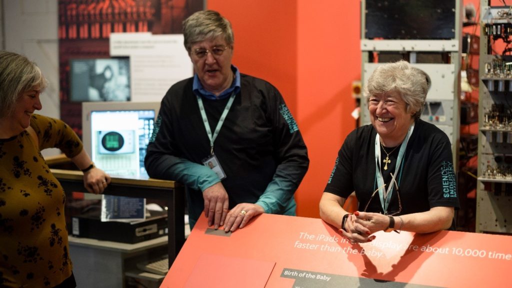 A man and a woman stood in front of an old computerr, talking to another woman