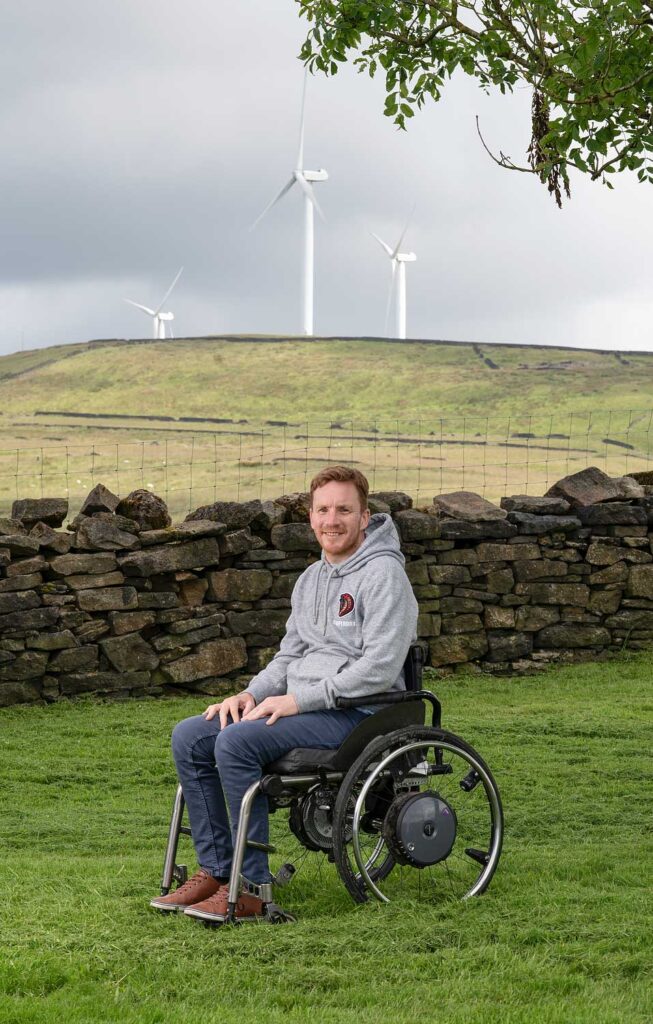 A man sat in a wheelchair in a field, with wind turbines in the background