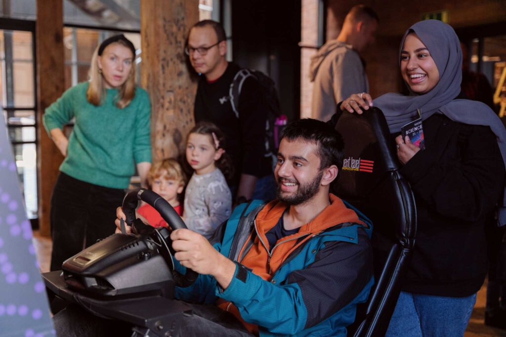 A man sat in a driving simulator with people stood behind him watching