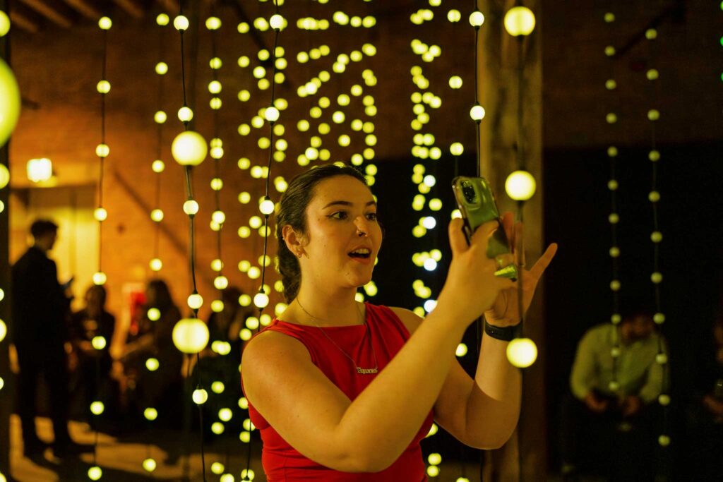 A woman in a red dress taking a photo of yellow spherical lights hanging from the ceiling of a warehouse