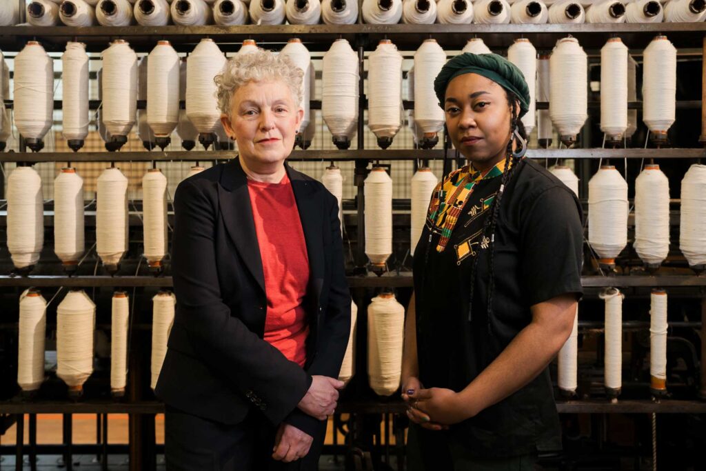Two women posing for a photograph in front of historic textile machinery